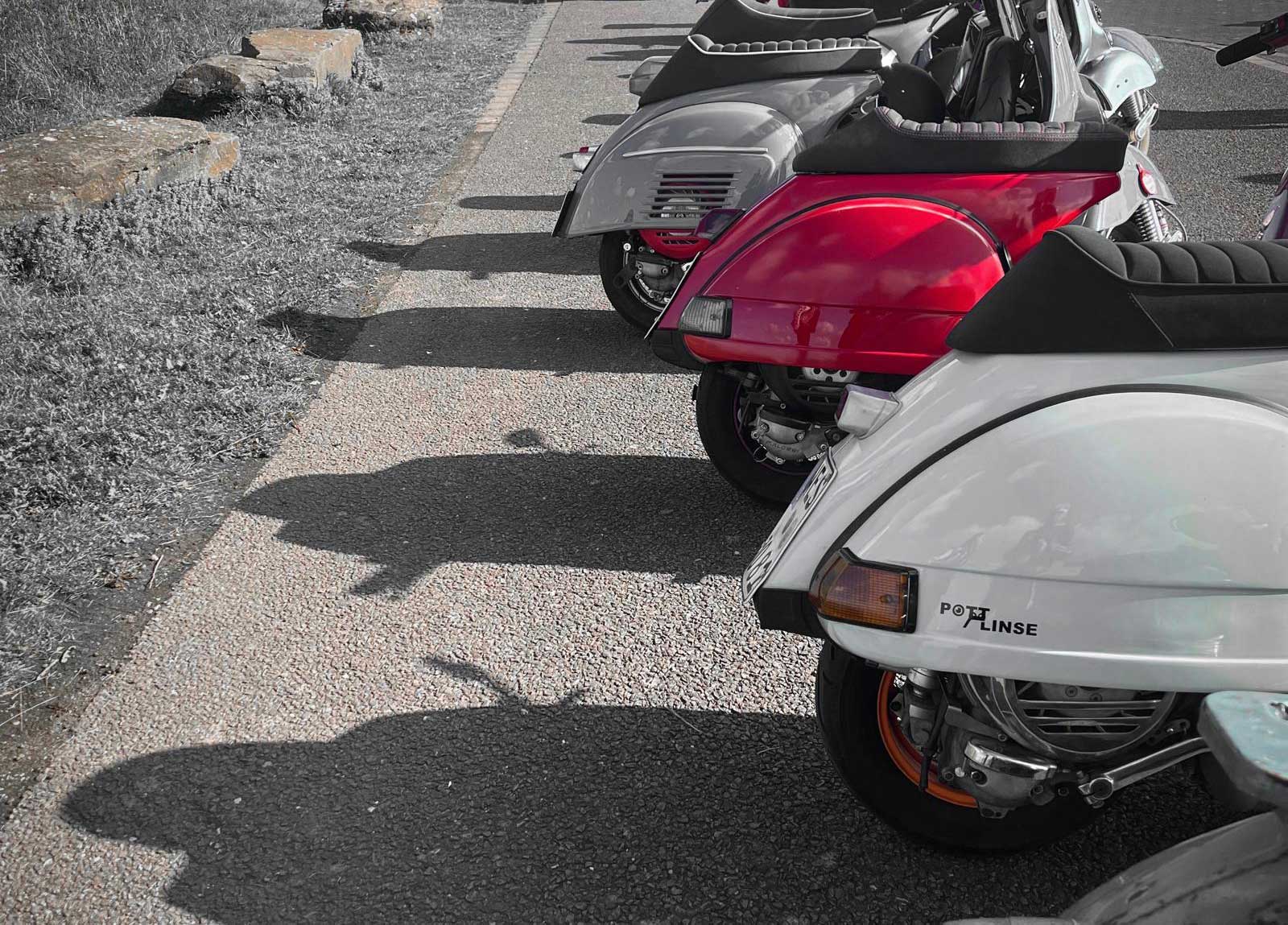 Reihe von motorrollern in verschiedenen farben, nebeneinander auf einem pflasterweg geparkt, mit gras und steinen links.